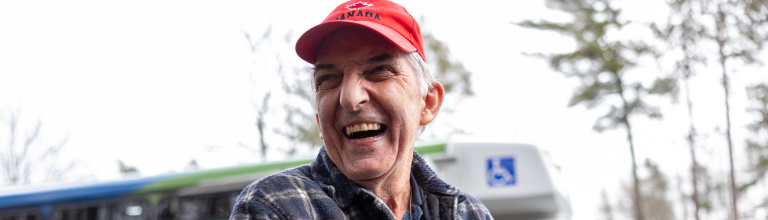 An older adult wearing a red hat and a flannel jacket smiling outside, with a transit bus is in the background.