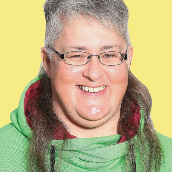 A smiling Caucasian woman with brown and grey hair that is short on top and long in the back, flowing to the side along the shoulders, wearing glasses and a green sweater with red plaid trim.