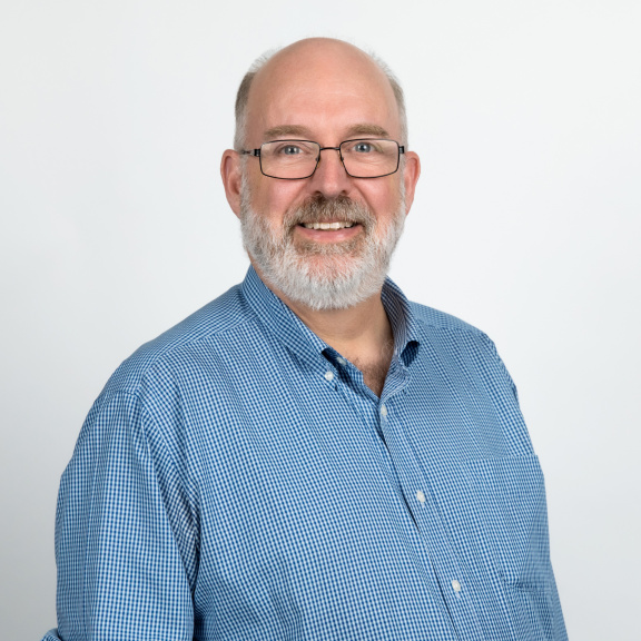 a white man with a beard, moustache and glasses in a blue shirt.
