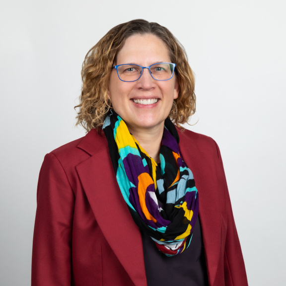 A smiling white woman with glasses and light brown hair, wearing a red jacket and a scarf.