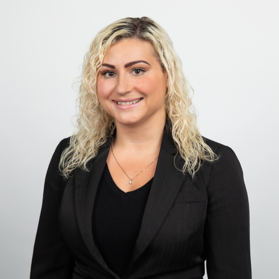 A smiling half Caucasian half Lebanese woman with curly blonde hair and dark brown roots, wearing a black shirt, blazer, and gold diamond necklace.