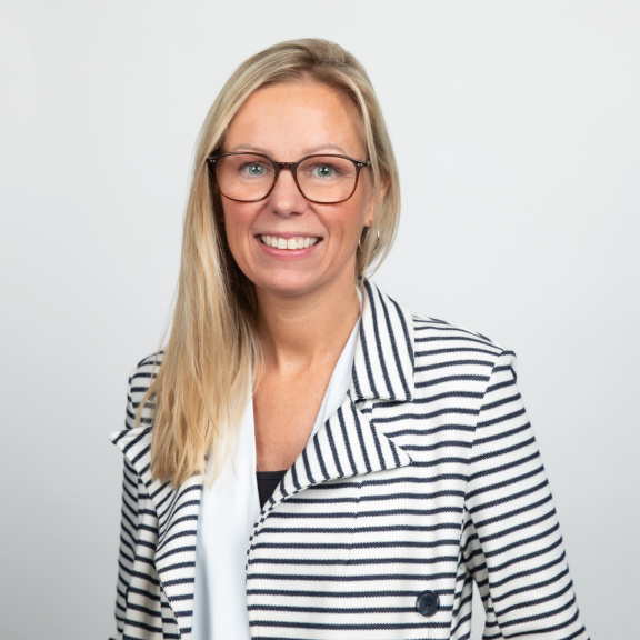 A smiling white woman with long blonde hair and glasses wearing a white and black striped blazar