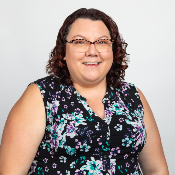 A smiling Caucasian woman with shoulder length dark brown curly hair wearing glasses and a sleeveless black shirt with a floral print.