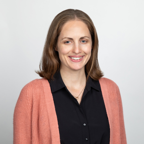 A white woman with shoulder length light brown hair.and blue eyes smiling and wearing a pink sweater over a black shirt.