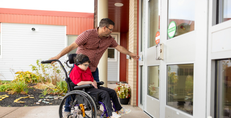 A middle-aged person wearing a flannel shirt is pushing a child in a wheelchair outside of a school. The person is pressing the automatic door push button to open the door.