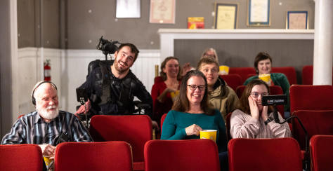 Group of people in sitting in a theatre watching a movie, a young person is using assistive technology for captioning and an older person is wearing headphones for described audio.