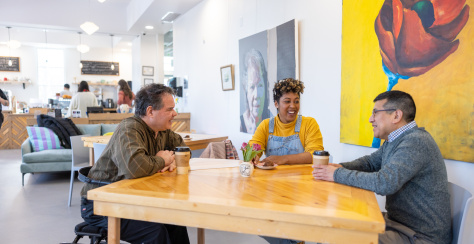 Three people sitting at a table in a bright café with large paintings in the background.