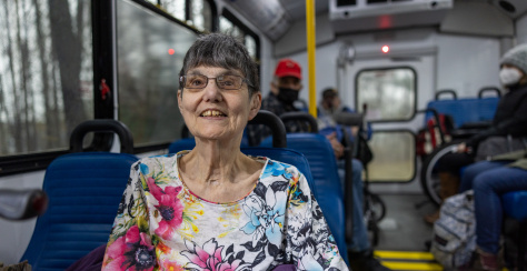 An older person wearing a floral shirt smiling while sitting on a bus.