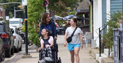 Three people walking down a sidewalk having a conversation. An adult is pushing a person in a powerchair and a younger person is walking beside them.