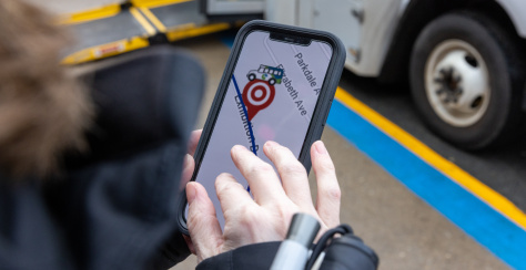 Photo of a person holding a smartphone with a map and “Target Location” marker, near a white vehicle and a painted curb, suggesting navigation.